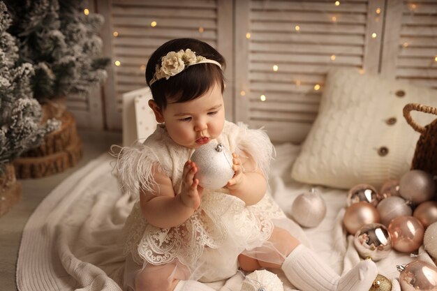 Une jolie fille à la peau foncée est assise sur une couverture tricotée blanche et joue avec des jouets d'arbre de Noël