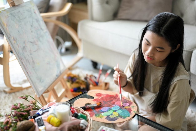 Jolie fille peignant ses œuvres d'art créatives sur un chevalet en toile et mélangeant la couleur acrylique sur une palette
