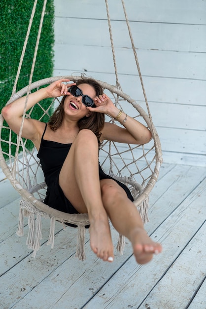 Jolie fille porte une élégante robe noire est assise dans une chaise berçante en plein air ou en studio