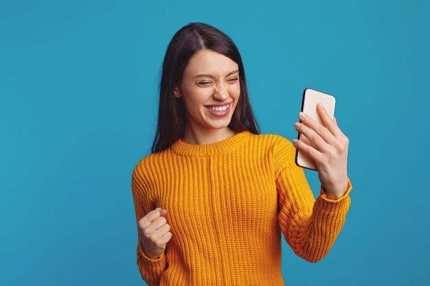 Jolie fille en pull jaune serrant le poing comme un gagnant lors de l'utilisation d'un mobile