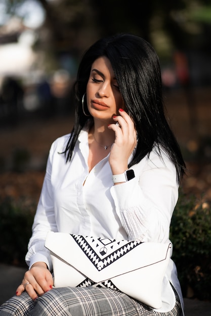 Jolie Fille Reposant Sur La Nature Le Week-end. Fille En Chemise Blanche Assise Dans Le Parc. Caucasienne Brune Jeune Femme Parlant Sur Smartphone à L'extérieur Seul. Technologies Et Gadgets.