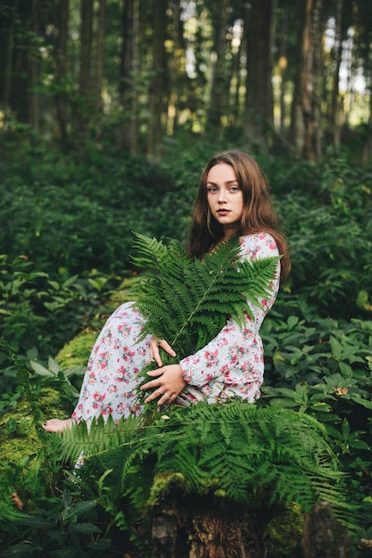 Une jolie fille en robe à fleurs est assise avec un bouquet de fougère dans la forêt.