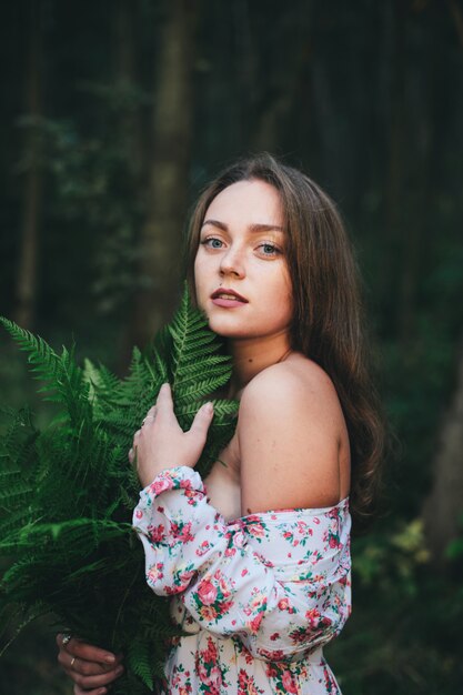 Une jolie fille en robe à fleurs est assise avec un bouquet de fougère dans la forêt.