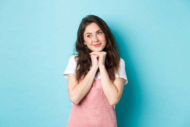 Jolie fille romantique avec une coiffure frisée, rêvant de quelque chose de beau, soupirant et souriant tendrement, l'air pensif à gauche, debout sur fond bleu