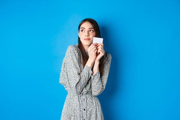 Jolie fille romantique en robe rêvant de faire du shopping, tenant une carte de crédit et regardant le logo avec un sourire tendre, debout sur le bleu.