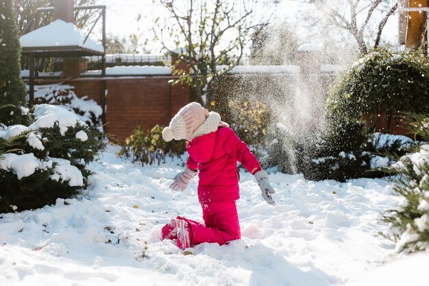 Jolie fille de sept ans en vêtements d'hiver jouant avec de la neige dans la cour d'une maison par une journée ensoleillée d'hiver.