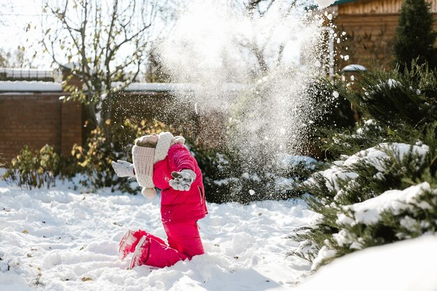 Jolie fille de sept ans en vêtements d'hiver jouant avec de la neige dans la cour d'une maison par une journée ensoleillée d'hiver.