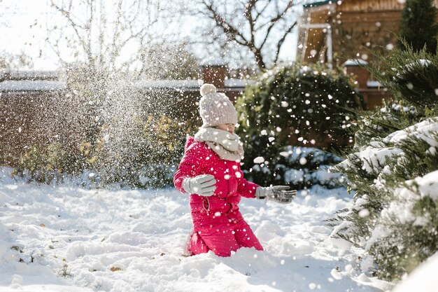 Jolie fille de sept ans en vêtements d'hiver jouant avec de la neige dans la cour d'une maison par une journée ensoleillée d'hiver.