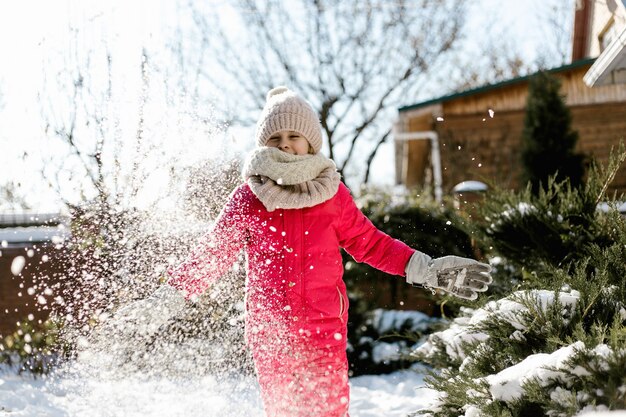 Jolie fille de sept ans en vêtements d'hiver jouant avec de la neige dans la cour d'une maison par une journée ensoleillée d'hiver.