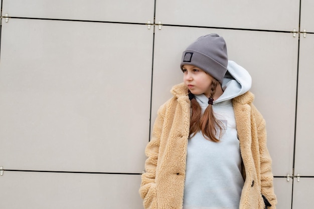 une jolie fille de six ans dans un manteau en fausse fourrure beige se tient contre un mur de la ville