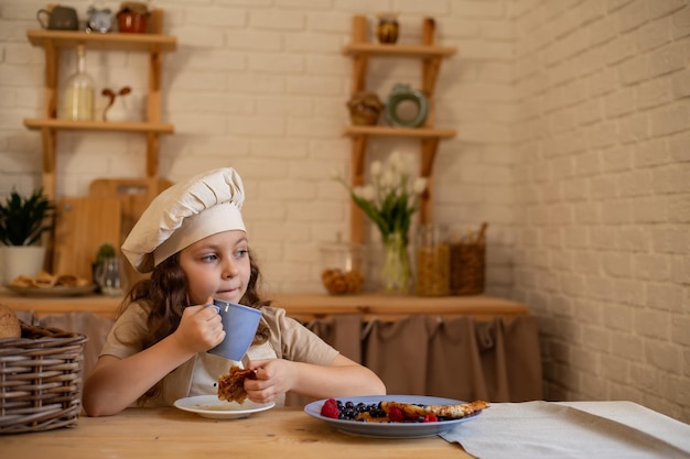 une jolie fille de six ans dans une toque et un tablier de chef est assise à une table en bois en train de manger des crêpes