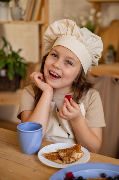 une jolie fille de six ans dans une toque et un tablier de chef est assise à une table en bois en train de manger des crêpes