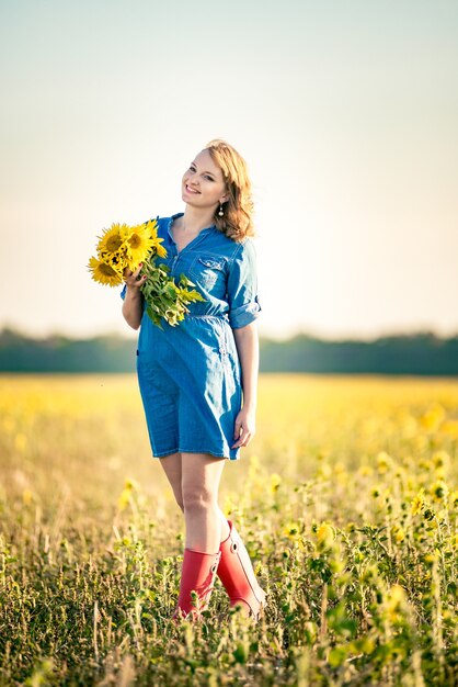 Jolie fille souriante aux cheveux roux vêtue d'une chemise en denim dans le champ plein de tournesols, bouquet de tournesols.