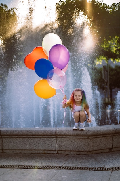 Jolie fille souriante caucasienne avec des cheveux teints colorés tenant un siège de ballons par fontaine Image wit