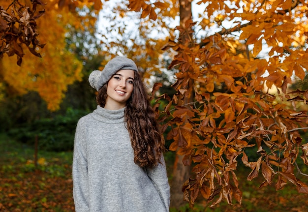 Jolie Fille Souriante élégante Aux Cheveux Bouclés Marchant Dans Le Parc Vêtu De La Mode Tendance Automne Gris Chaud, Portant Un Béret.