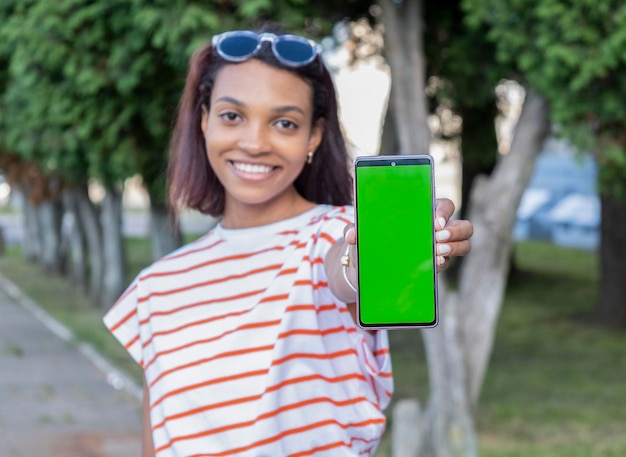 Jolie fille souriante à la peau foncée en t-shirt blanc de base montrant un écran vert vierge de son smartphone à la caméra Maquette de l'espace de copie pour votre logo texte ou votre interface