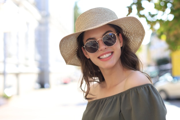 Jolie fille souriante portant des lunettes de soleil et un chapeau d'été et regardant la caméra en marchant dans la rue de la ville.