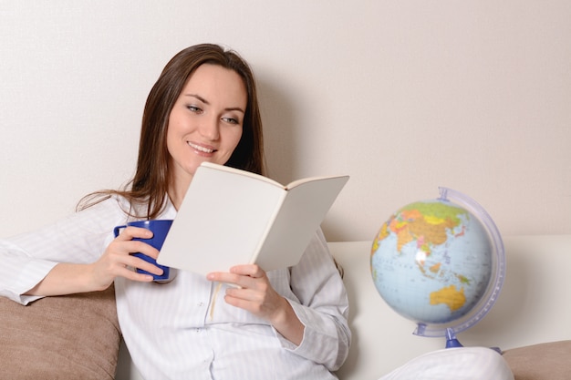 Jolie fille souriante avec une tasse bleue et le journal, assise sur le canapé dans la chambre et lit