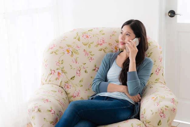 jolie fille souriante utilisant un téléphone portable appelant un ami bavardant le week-end lorsqu'elle est assise devant le canapé de la fenêtre pour se détendre.