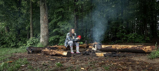 Une jolie fille avec une tasse à la main est assise sur une bûche et se réchauffe près d'un feu dans la forêt.