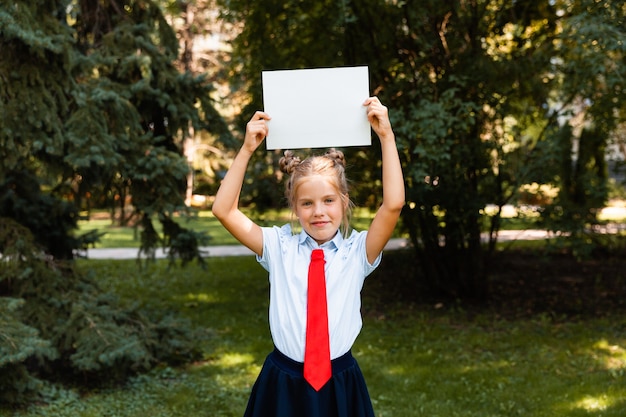 Jolie fille tenant un drap blanc. Jeune fille blonde avec une feuille de papier blanche. La petite écolière tient une feuille blanche.