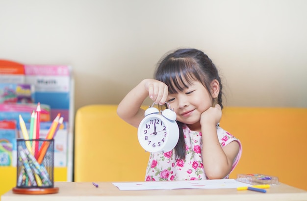 Une jolie fille tenant une horloge alors qu'elle est assise à la table à la maison.