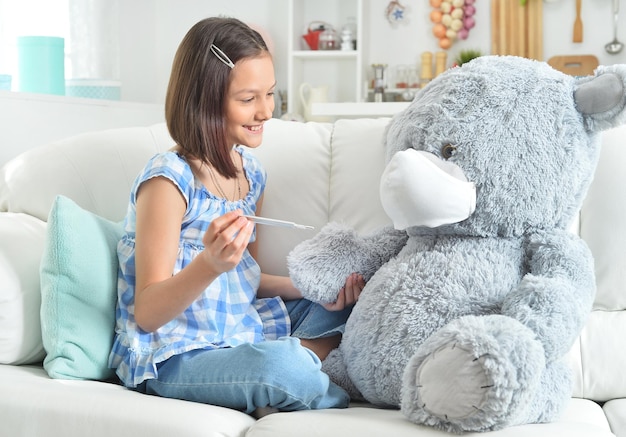 Jolie fille avec thermomètre jouant avec un ours en peluche dans un masque facial