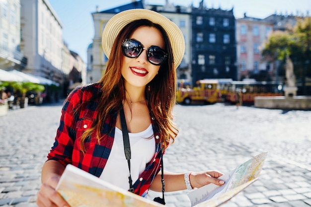 Jolie fille touristique aux cheveux bruns portant un chapeau, des lunettes de soleil et une chemise rouge, tenant une carte dans la vieille ville européenne et souriant, voyageant.