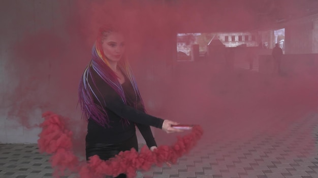 Jolie fille avec des tresses multicolores et du maquillage dans une robe noire. Posant se cachant derrière la fumée artificielle rouge dans le contexte d'un bâtiment dans une ville printanière.