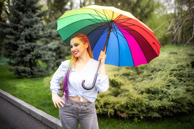 Une jolie fille avec des tresses multicolores et un maquillage lumineux dans une chemise bleuâtre posant avec un parapluie arc-en-ciel dans le contexte d'un parc printanier en fleurs