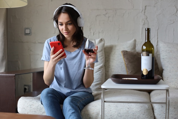 Jolie fille utilisant son smartphone sur un canapé à la maison dans le salon. Écouter de la musique, boire du vin rouge, se détendre après une dure semaine de travail.