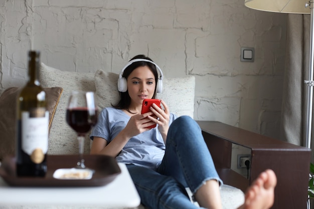 Jolie fille utilisant son smartphone sur un canapé à la maison dans le salon. Écouter de la musique, boire du vin rouge, se détendre après une dure semaine de travail.