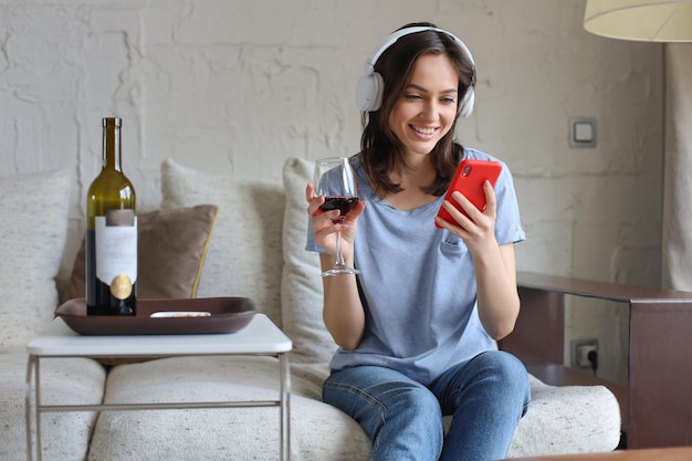 Jolie fille utilisant son smartphone sur un canapé à la maison dans le salon Écouter de la musique en buvant du vin rouge se détendre après une dure semaine de travail