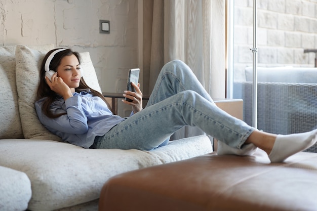 Jolie fille utilisant son smartphone sur un canapé à la maison dans le salon. Écouter de la musique, détente après une dure semaine de travail