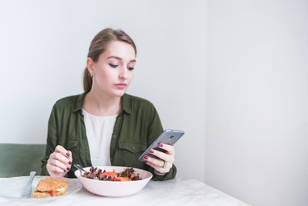 jolie fille utilise smearphone tout en mangeant au restaurant. femme mange de la salade et regarde smartphone