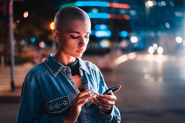 Jolie fille avec des vêtements élégants tenant le smartphone à l'extérieur le soir, ville illuminée