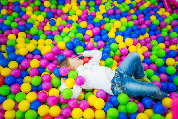 Jolie fille en vêtements pour enfants joue dans la piscine avec des boules en plastique brillant de différentes couleurs