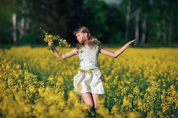 Une jolie fille vêtue d'une robe blanche traverse un champ de colza avec un bouquet dans les mains et rit