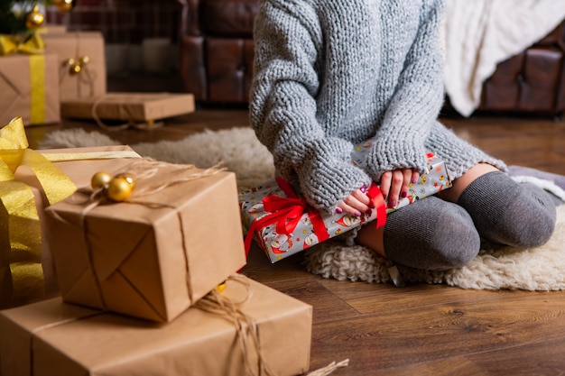 Une jolie fille vêtue d'une robe chaude s'assoit sur un tapis et noue un cadeau avec un ruban pour le Nouvel An près d'autres boîtes contenant des cadeaux dans la chambre