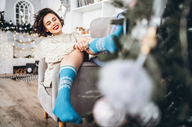 Jolie et heureuse brune assise en pull chaud sur la chaise avec des lumières de Noël derrière