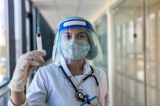 Jolie infirmière en uniforme et écran facial tenant la seringue et en attente de vaccination en clinique