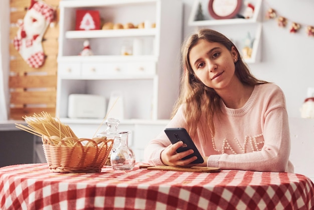 Jolie jeune adolescente avec téléphone dans les mains est assise près de la table dans la cuisine pendant la journée.