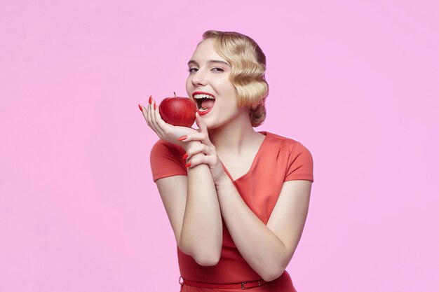 Jolie jeune blonde avec une coiffure rétro est sur le point de mordre une pomme rouge