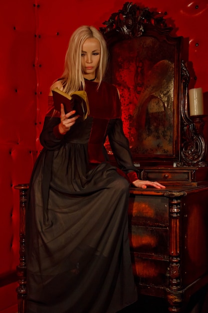 Jolie jeune blonde sexy en robe gothique à l'intérieur de la salle rouge médiévale avec un vieux miroir lit la Bible et montre des émotions. Image de la reine d'horreur d'Halloween. Copiez l'espace pour le texte ou le logo