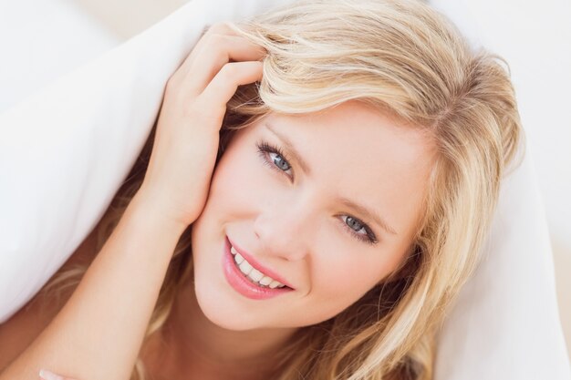 Jolie jeune blonde souriante à la caméra de sous la couette