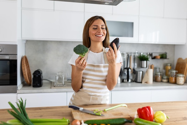 Une jolie jeune brune heureuse de bonne humeur prépare une salade végétalienne fraîche pour une vie saine dans la cuisine de sa maison