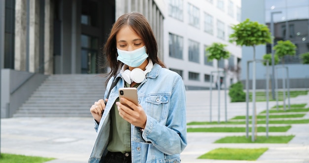Jolie jeune étudiante élégante caucasienne en masque médical et avec un message texto sac à dos sur téléphone mobile. Belle femme tapant et faisant défiler sur smartphone. Concept de pandémie. En plein air.
