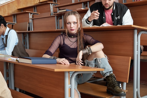 Jolie jeune étudiante en tenue décontractée vous regarde assis près d'un long bureau en bois dans une salle de conférence parmi des coéquipiers masculins interculturels