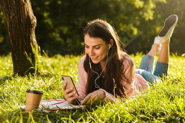 Jolie jeune étudiante utilisant un téléphone portable tout en étant allongée sur une herbe dans le parc, écoutant de la musique avec des écouteurs