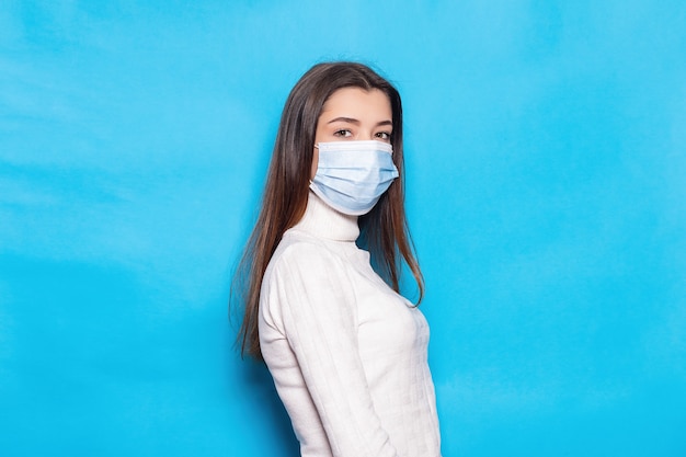 Jolie jeune femme de 20 ans dans un masque facial à l'abri du virus du coronavirus covid-19 pendant la quarantaine pandémique tenant les mains croisées isolées sur fond de couleur bleu vif portrait en studio